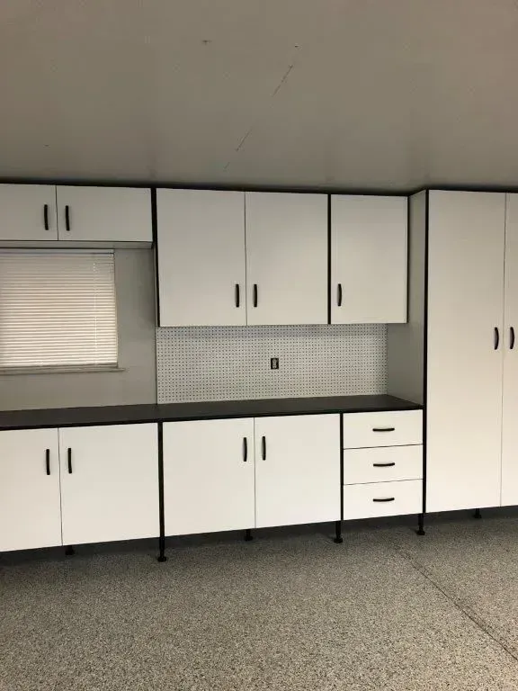White and black storage cabinets in a garage. Cabinets have black handles and trim, with a black countertop and a tiled backsplash.