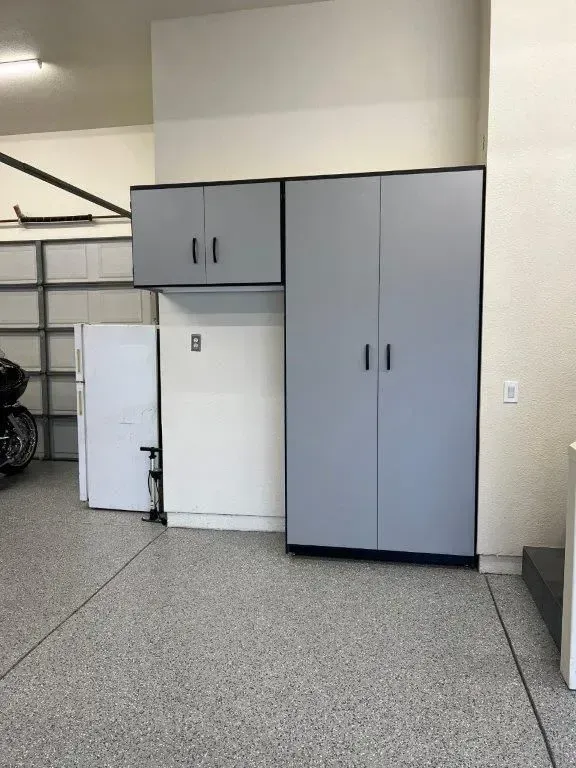 Garage storage cabinets, gray doors, and overhead cabinet against a white wall with a refrigerator.