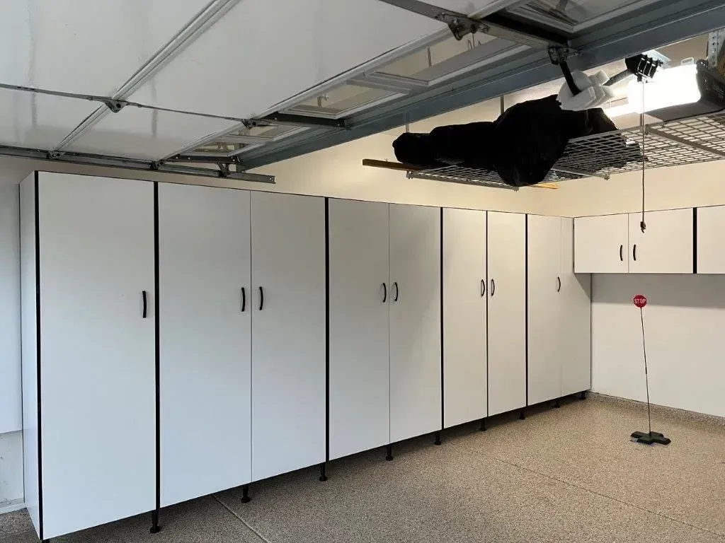 White storage cabinets line a garage wall; an overhead storage rack is above.