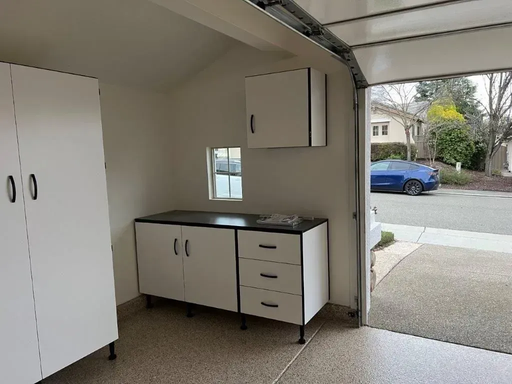 White garage cabinets, workbench, and tall cabinet in a garage with the door open to a street with a blue car.