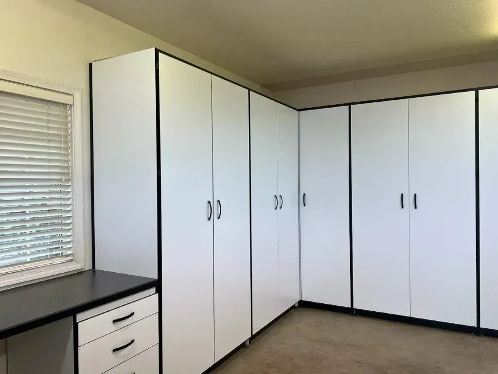 White storage cabinets with black trim in a corner room, next to a window and desk.