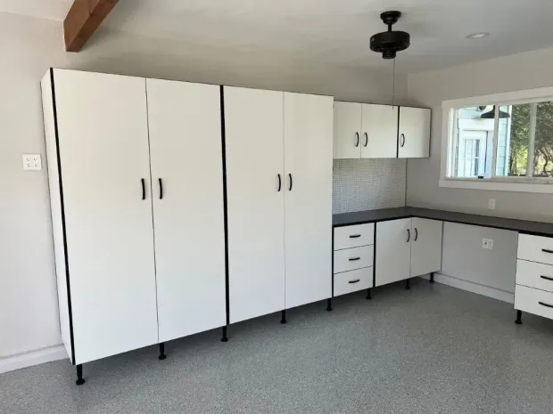 White garage storage cabinets with black accents and hardware against a light gray wall and speckled floor.