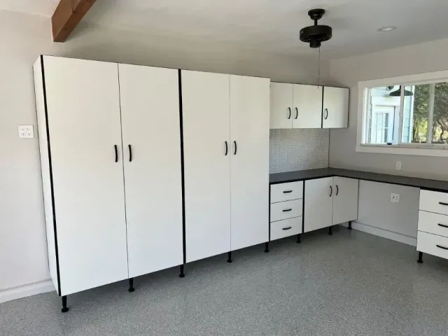 White garage storage cabinets with black accents and hardware against a light gray wall and speckled floor.