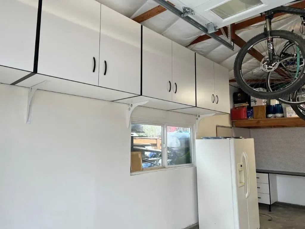 White overhead cabinets installed in a garage, with a window and refrigerator below.