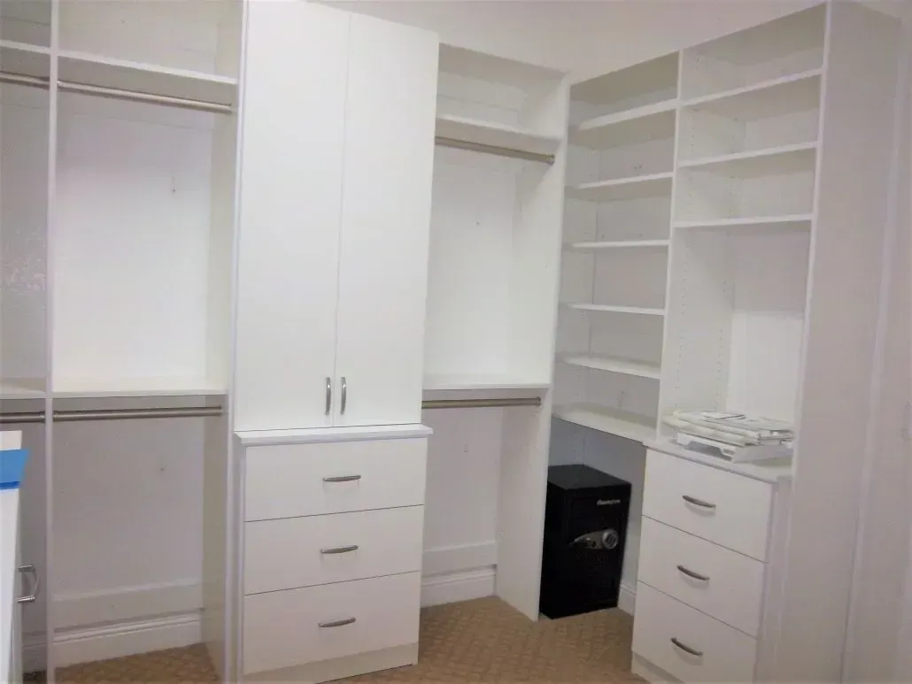 Walk-in closet with white shelving, cabinets, and drawers. A small safe is visible in a corner.
