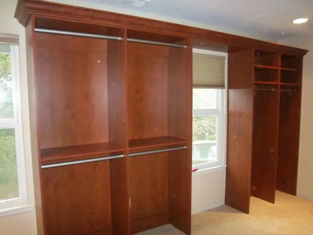 Wooden built-in closet with hanging rods and shelves, brown finish, in front of a window.