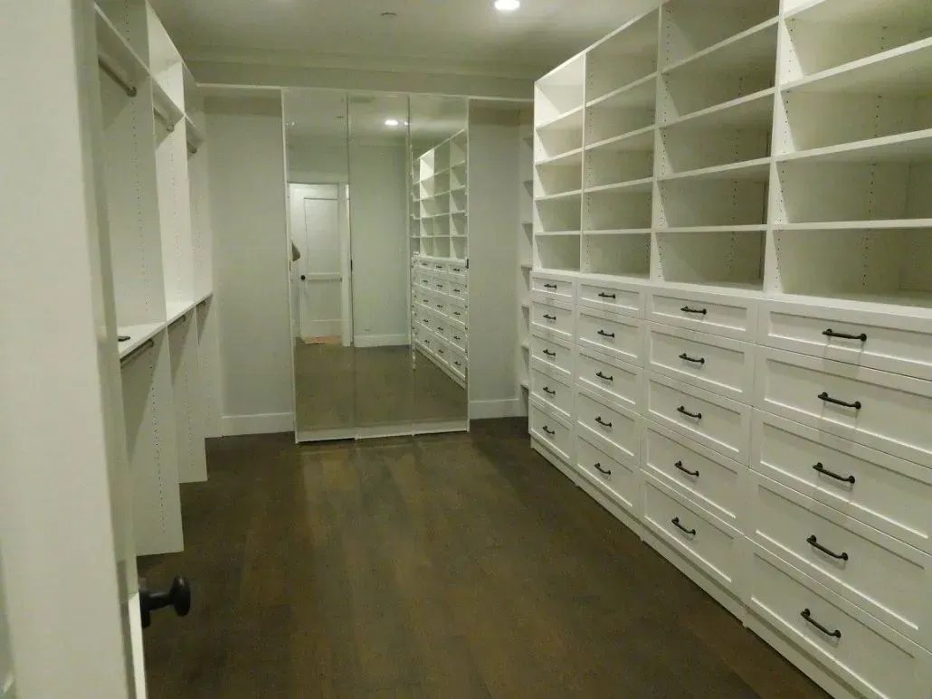 Spacious white walk-in closet with shelves, drawers, and a full-length mirror, dark wood floor.
