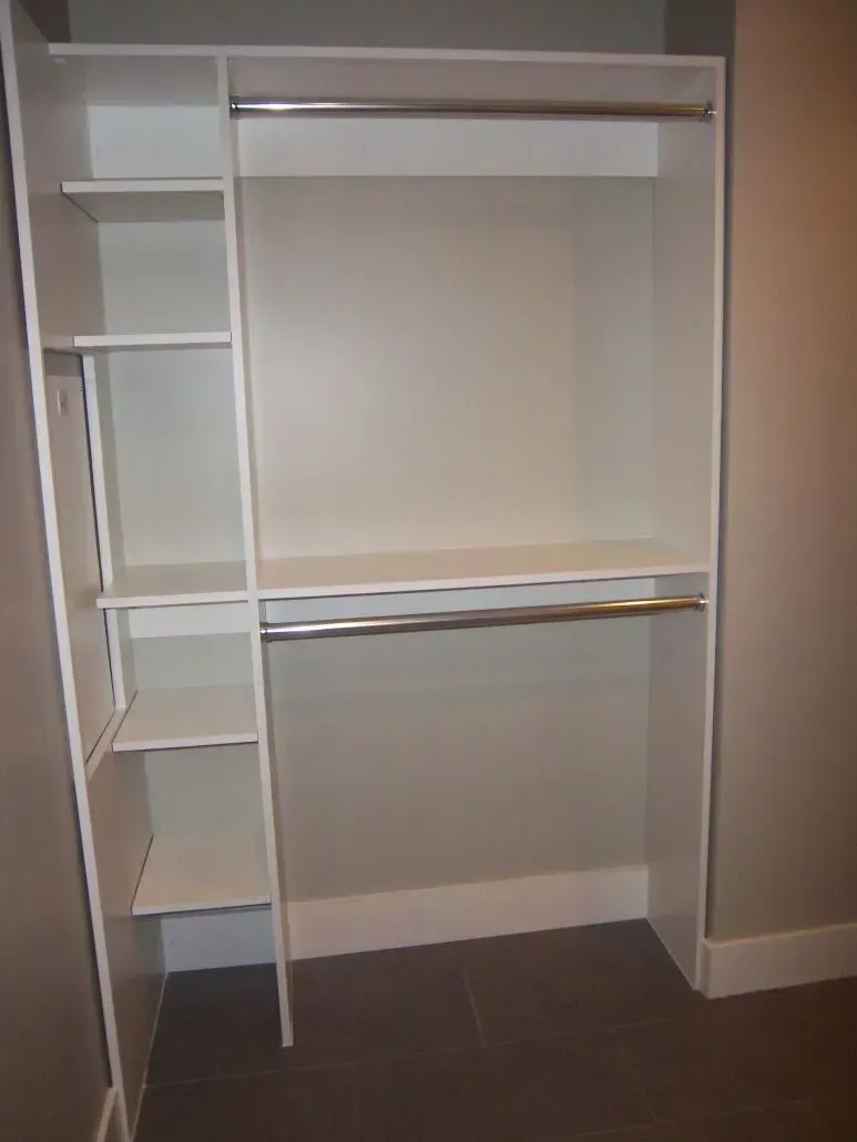 White built-in closet with shelves and rods, inside a room with beige walls and a dark floor.