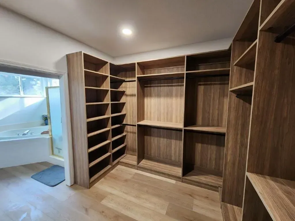 Walk-in closet with shelves and hanging rods. Light wood cabinetry, light wood floors, and a bathroom visible.