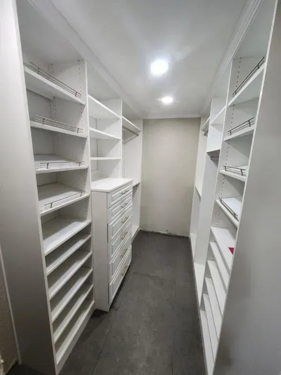 White walk-in closet with shelves, drawers, and hanging rods. Dark floor, plain walls, overhead lighting.