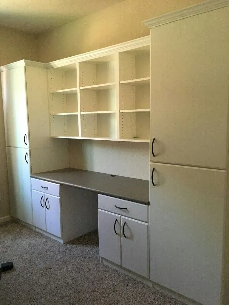 White built-in desk with cabinets and open shelves on carpeted floor against a beige wall.