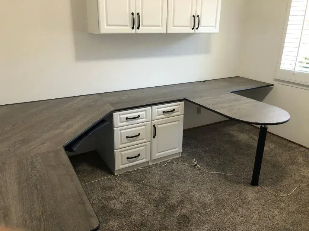 L-shaped desk with drawers and cabinets, under wall-mounted cabinets, on carpet, near a window.