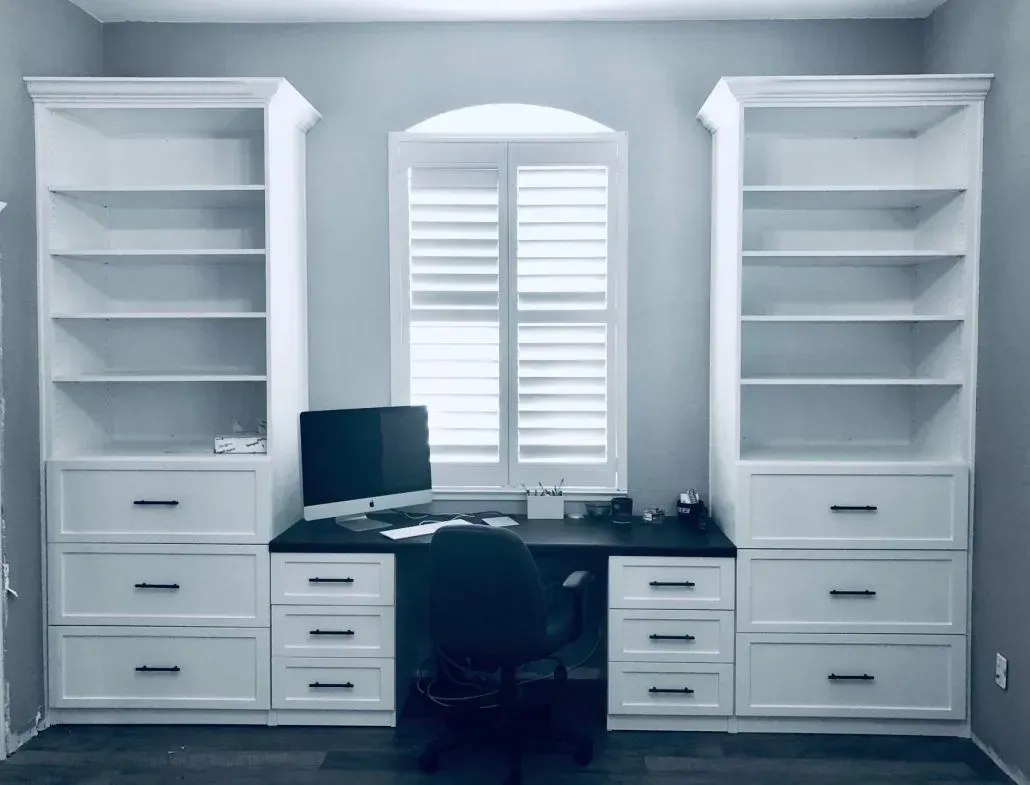 Home office with built-in white bookshelves, desk, and window with shutters. Black computer, chair, and desk top.