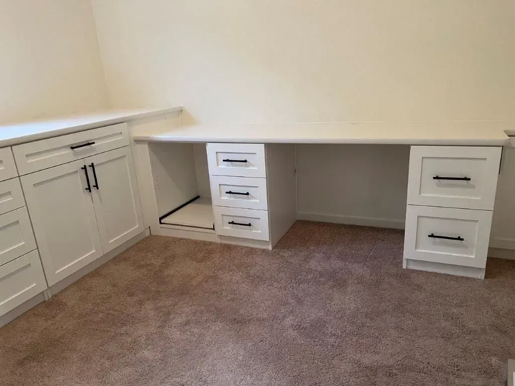 Built-in white desk with drawers and cabinets in a room with carpet. Black hardware on drawers and doors.