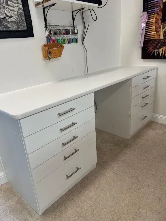 White desk with drawers against a wall. Sewing supplies and art are visible.