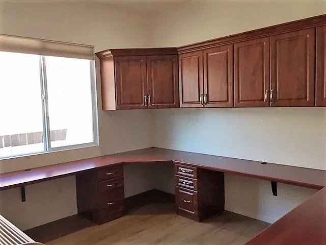 L-shaped home office with brown wood desk and overhead cabinets, next to a window.