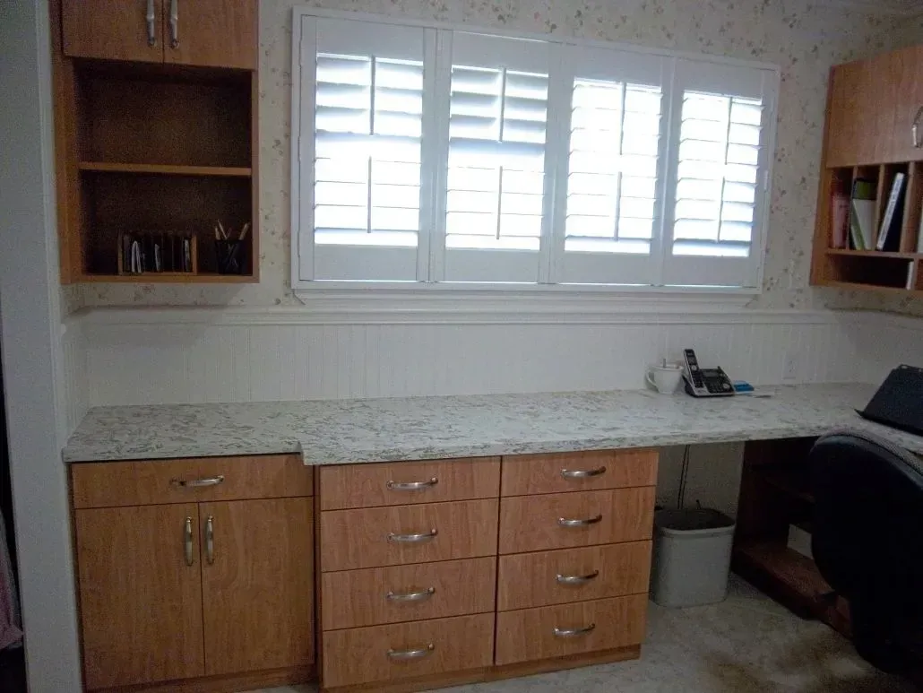 Built-in desk with cabinets and drawers, beneath a window with white shutters. Wood shelves on either side.