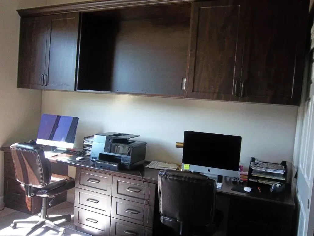 Home office with two computers on a desk, built-in cabinets above, and two office chairs.