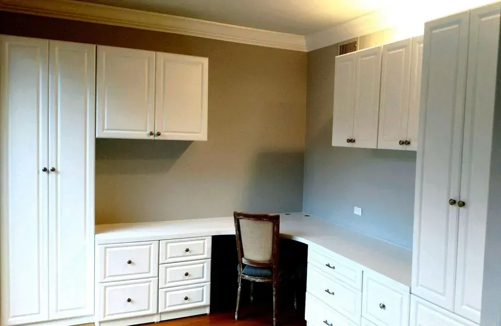 White built-in office cabinetry, including a desk, drawers, and overhead cabinets, with a chair in the center.