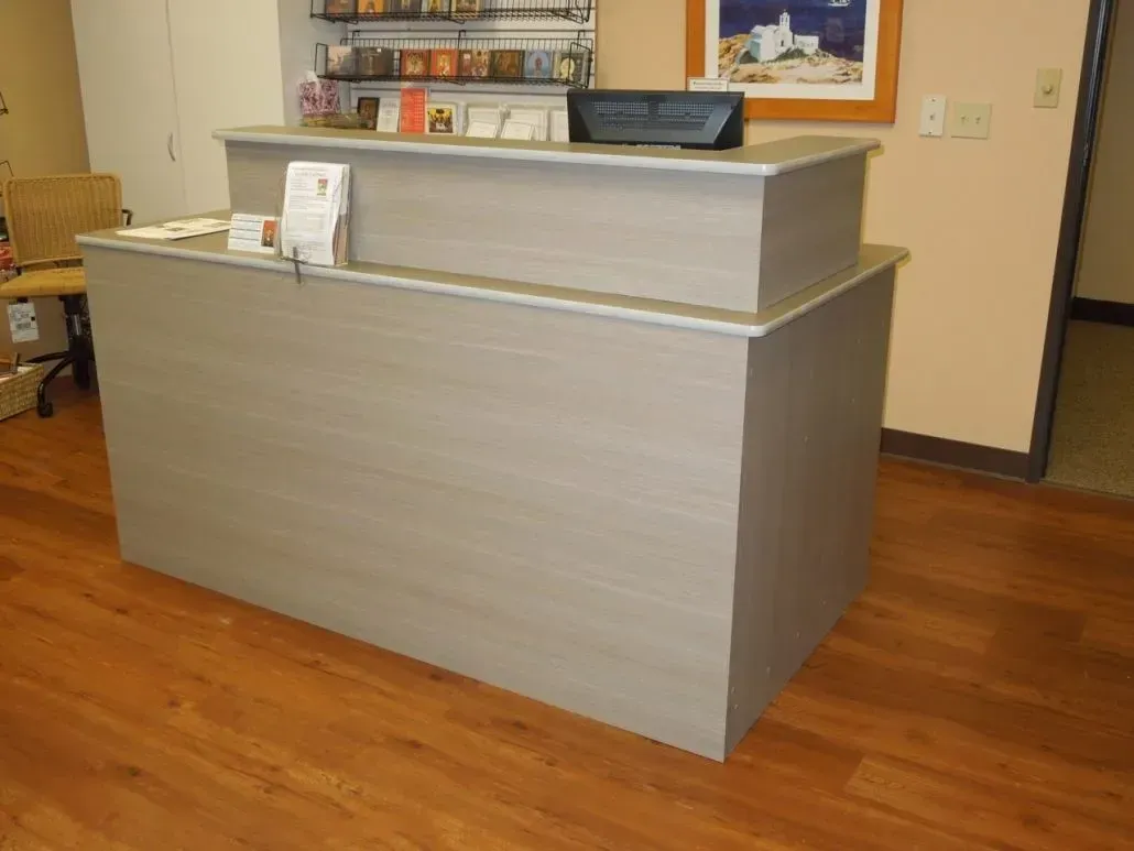 Reception desk in a clinic or office setting; light wood-grain finish, two tiers, on a wooden floor.