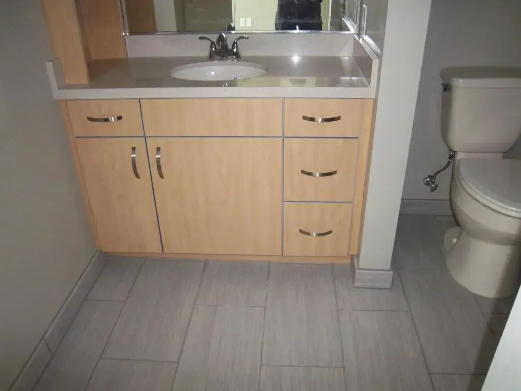 Bathroom vanity with sink, light wood cabinets, and gray tile floor. Toilet is beside the vanity.