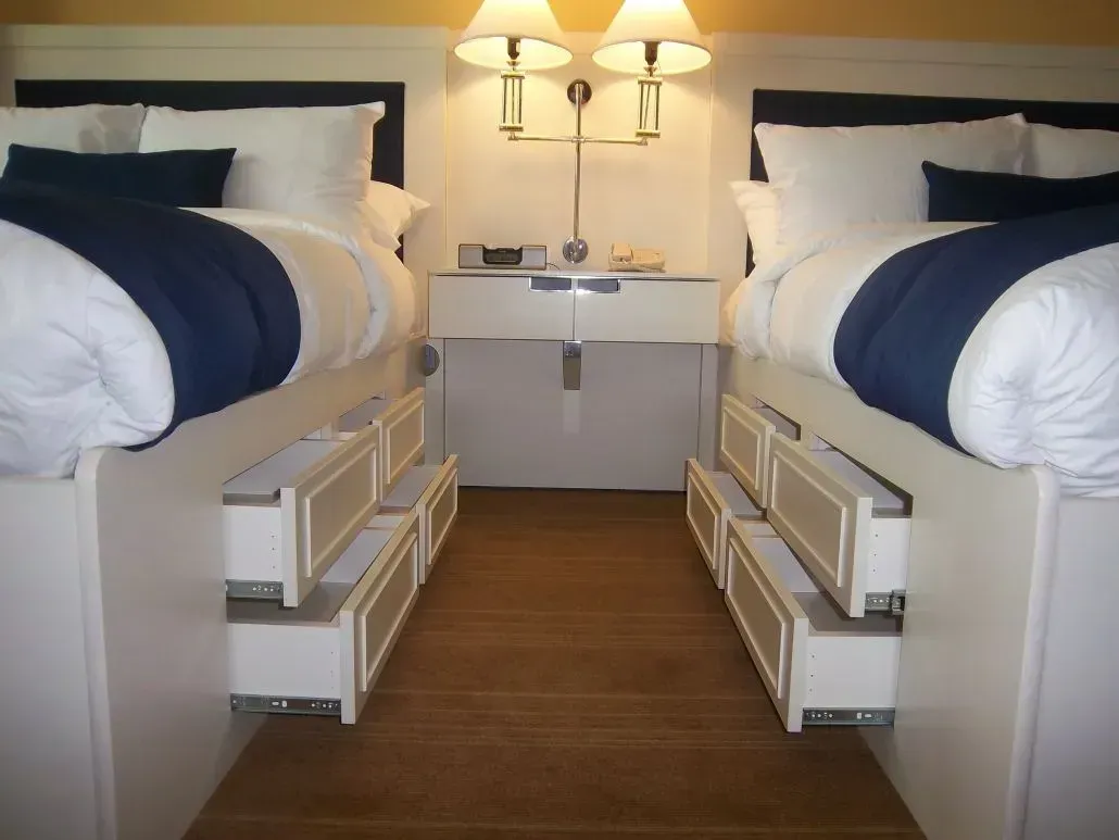 Two beds with blue accents and drawers beneath them, bedside table between them, two lamps.