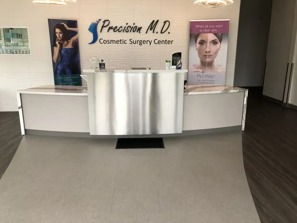 Reception area of Precision M.D. Cosmetic Surgery Center with a curved white desk and promotional banners.
