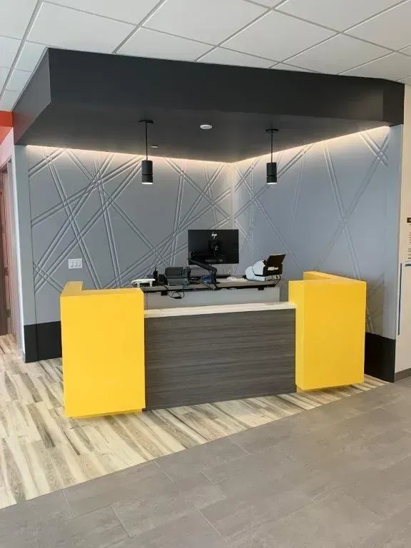 Reception desk with yellow panels, dark wood counter, gray wall with geometric pattern, two pendant lights.