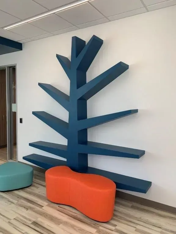 Blue tree-shaped bookshelf against white wall; orange and teal ottomans on the floor.