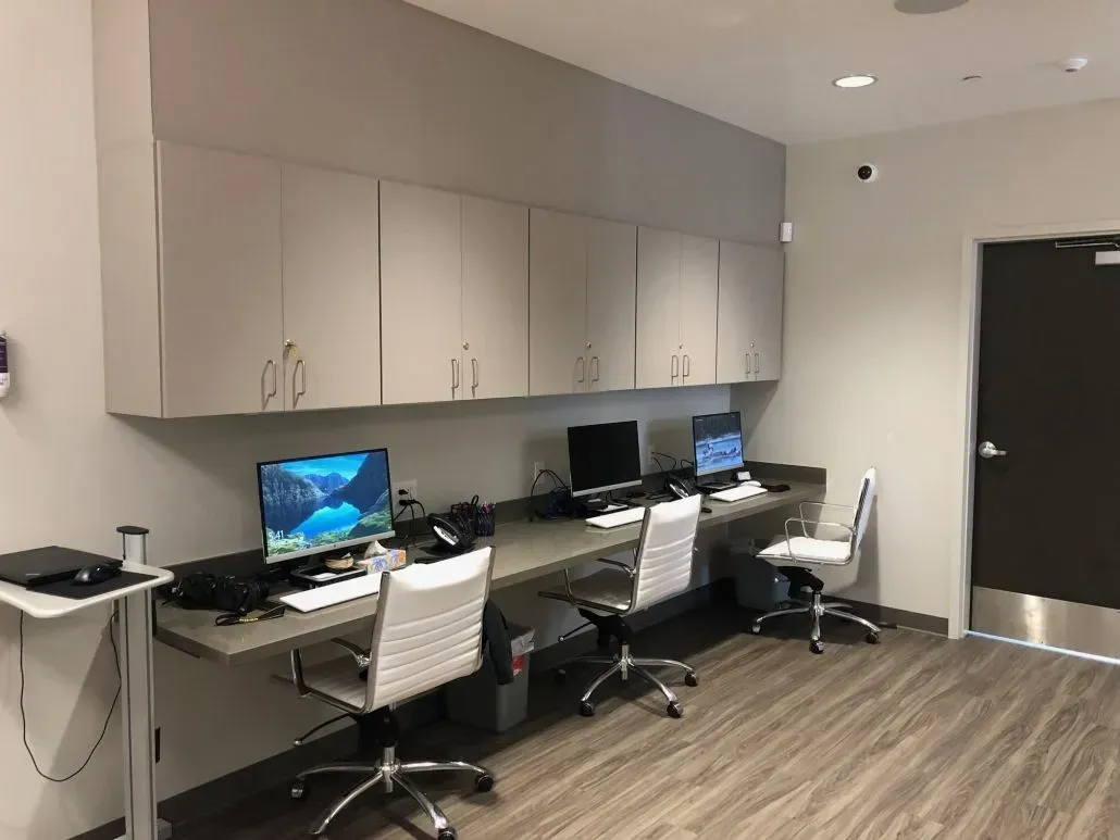 Computer workstations in a neutral-toned office with overhead storage, wood-look flooring, and a dark door.