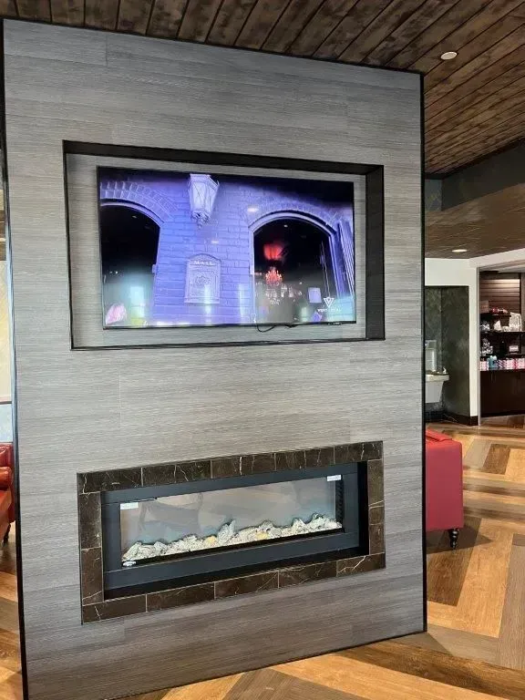 A gray wall with a mounted TV and a fireplace below. The TV displays a building, and the fireplace has a faux flame.
