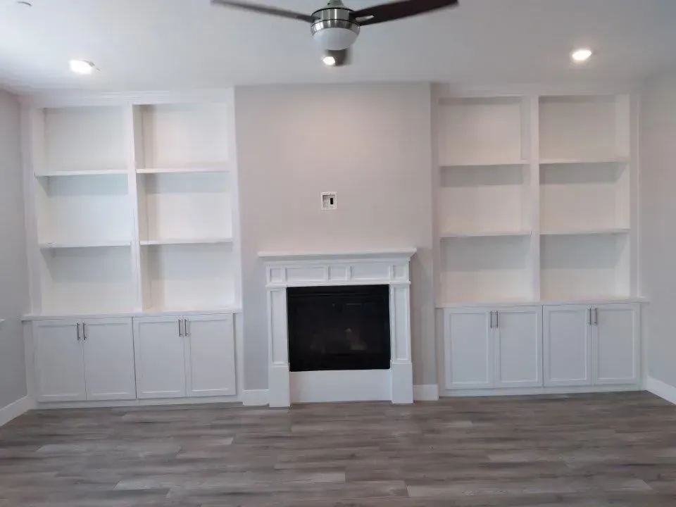 White built-in bookshelves frame a fireplace in a modern living room. Gray flooring and ceiling fan.