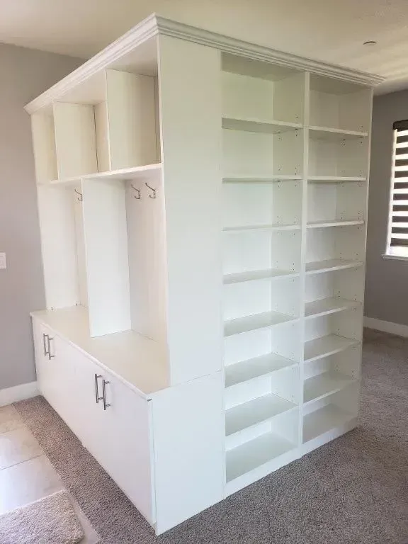 White built-in storage unit with shelves, hooks, and bench seating, set against a neutral wall and carpeted floor.