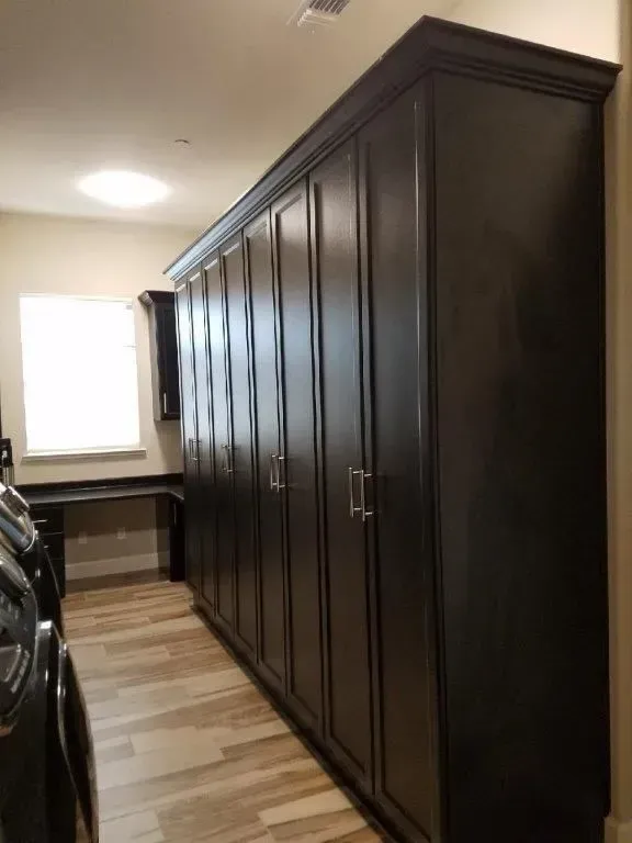 Dark brown storage cabinets line a laundry room wall. Light wood-look flooring and a small desk under a window are visible.