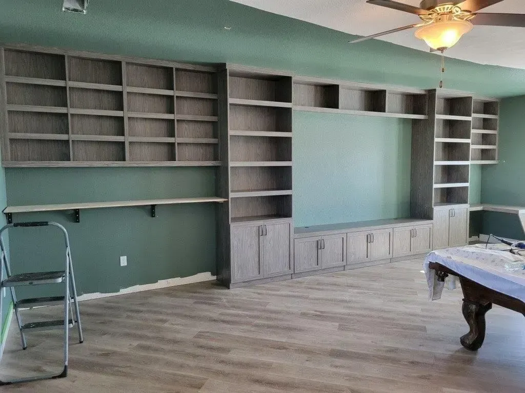 Built-in wooden shelving unit, desk and cabinet in room with teal walls, wood floor, and ceiling fan.