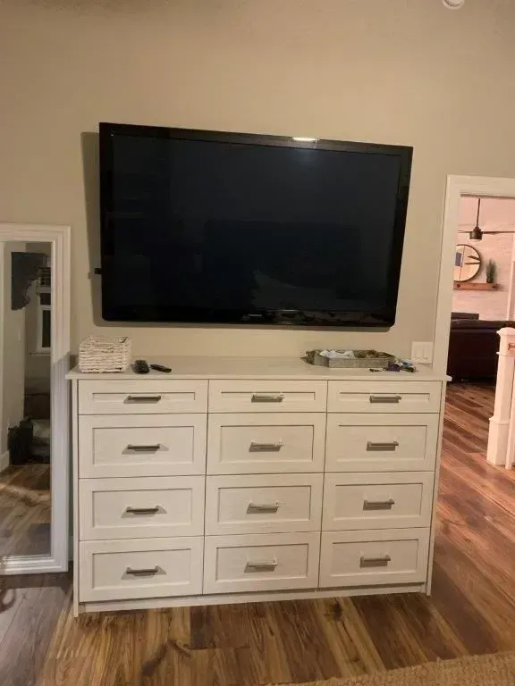 A TV mounted above a white dresser with nine drawers in a room with wood flooring.