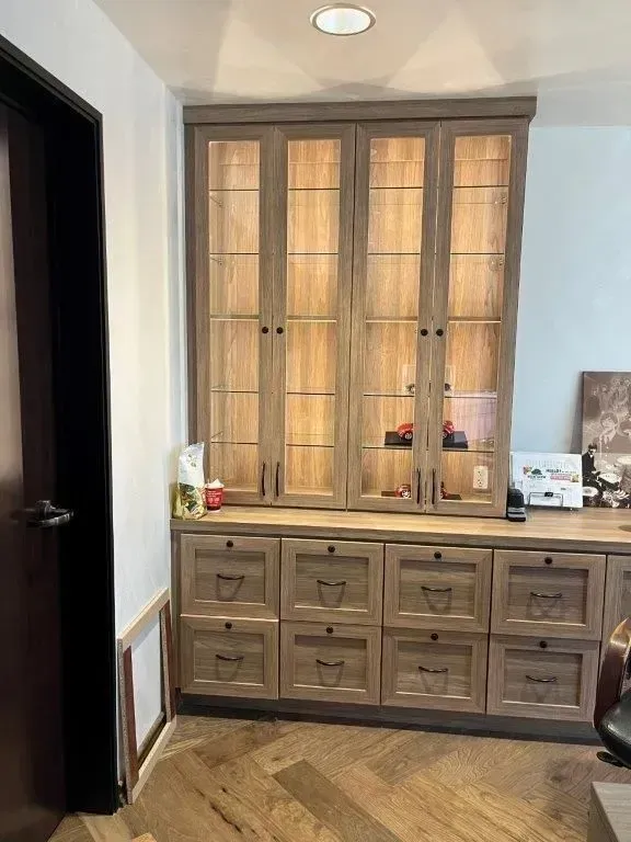 Wooden built-in cabinet with glass doors on top, drawers below. Lit interior, dark wood door on the left, wood floor.