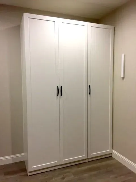 White three-door wardrobe against a beige wall, black handles.