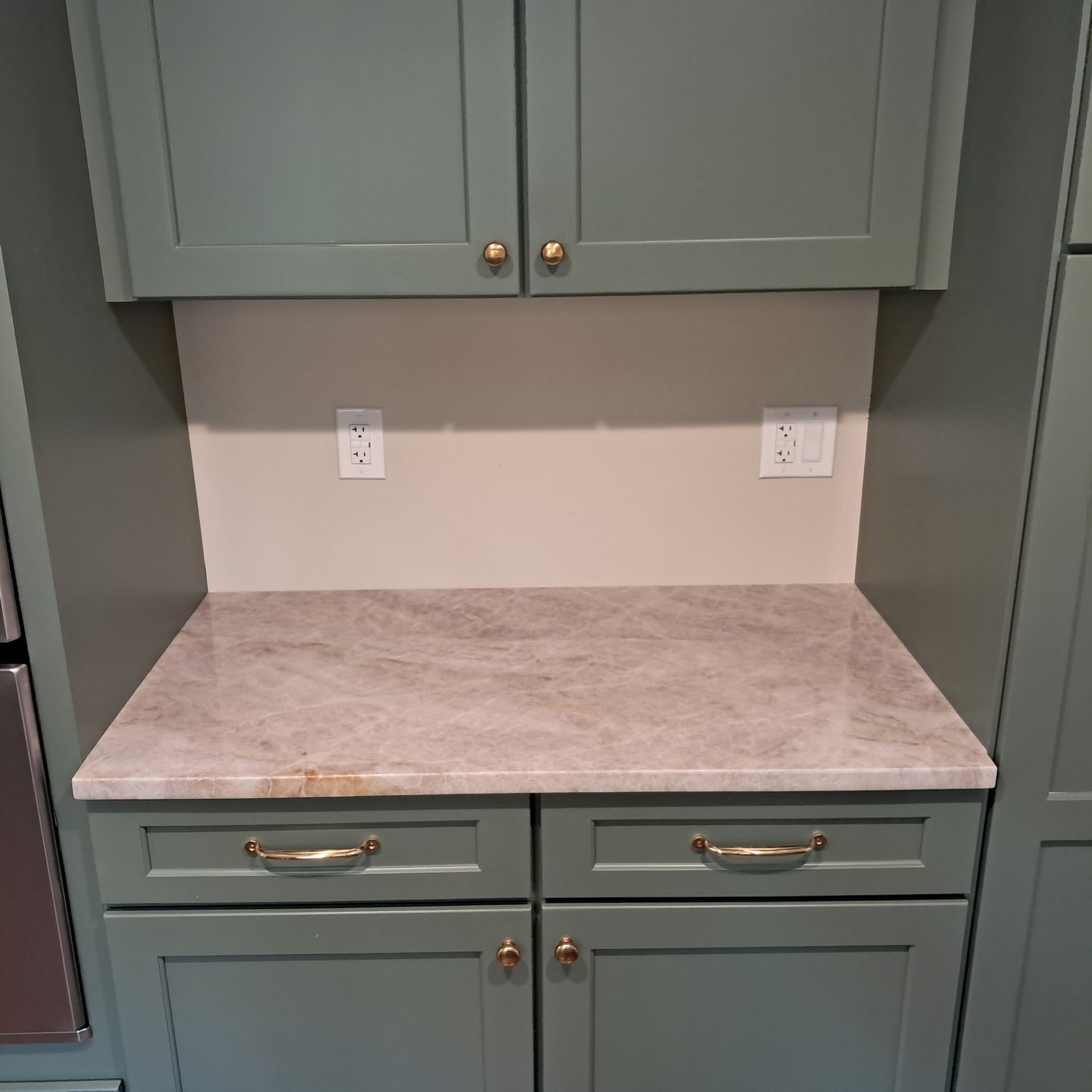 Green kitchen cabinets with beige countertop and backsplash, gold hardware, and electrical outlets.