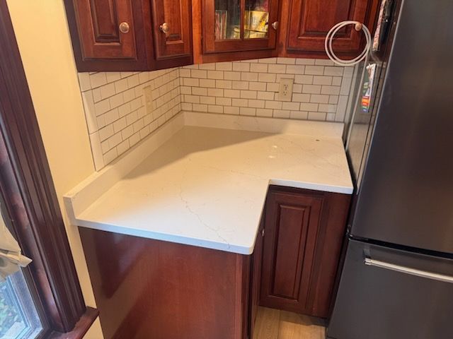 Kitchen corner with white countertop, subway tile backsplash, and dark wooden cabinets. A refrigerator is on the right.