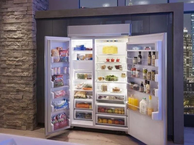 Open refrigerator filled with food, lit up. Stone wall and city view in background.
