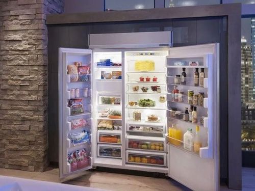 Open refrigerator filled with food, lit up. Stone wall and city view in background.