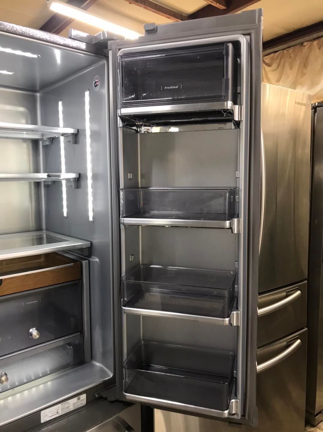 Open refrigerator door, revealing interior shelves and side storage. Stainless steel finish, bright interior light.