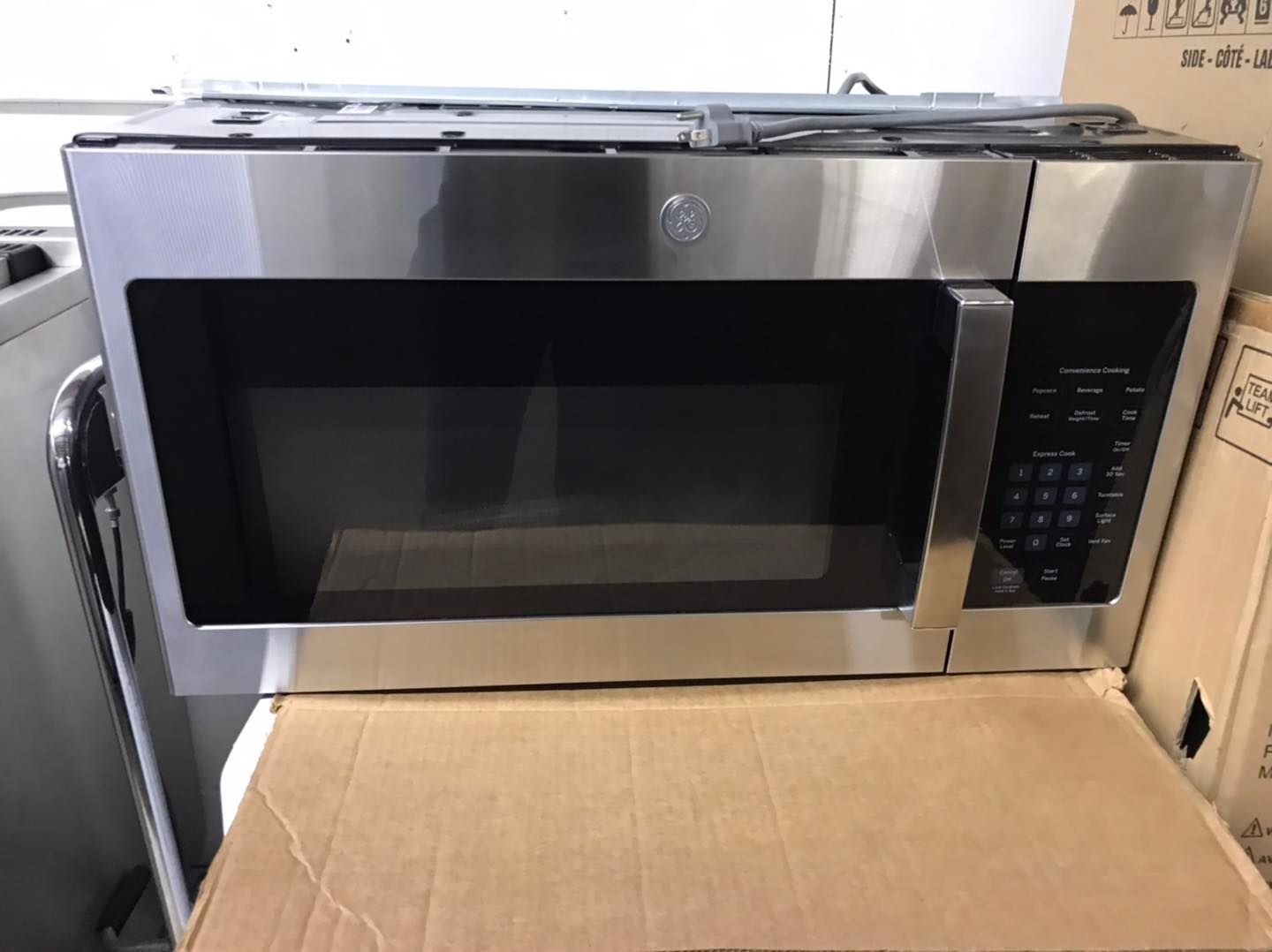 Stainless steel GE over-the-range microwave oven, on a cardboard box. Digital keypad and door handle on the right.