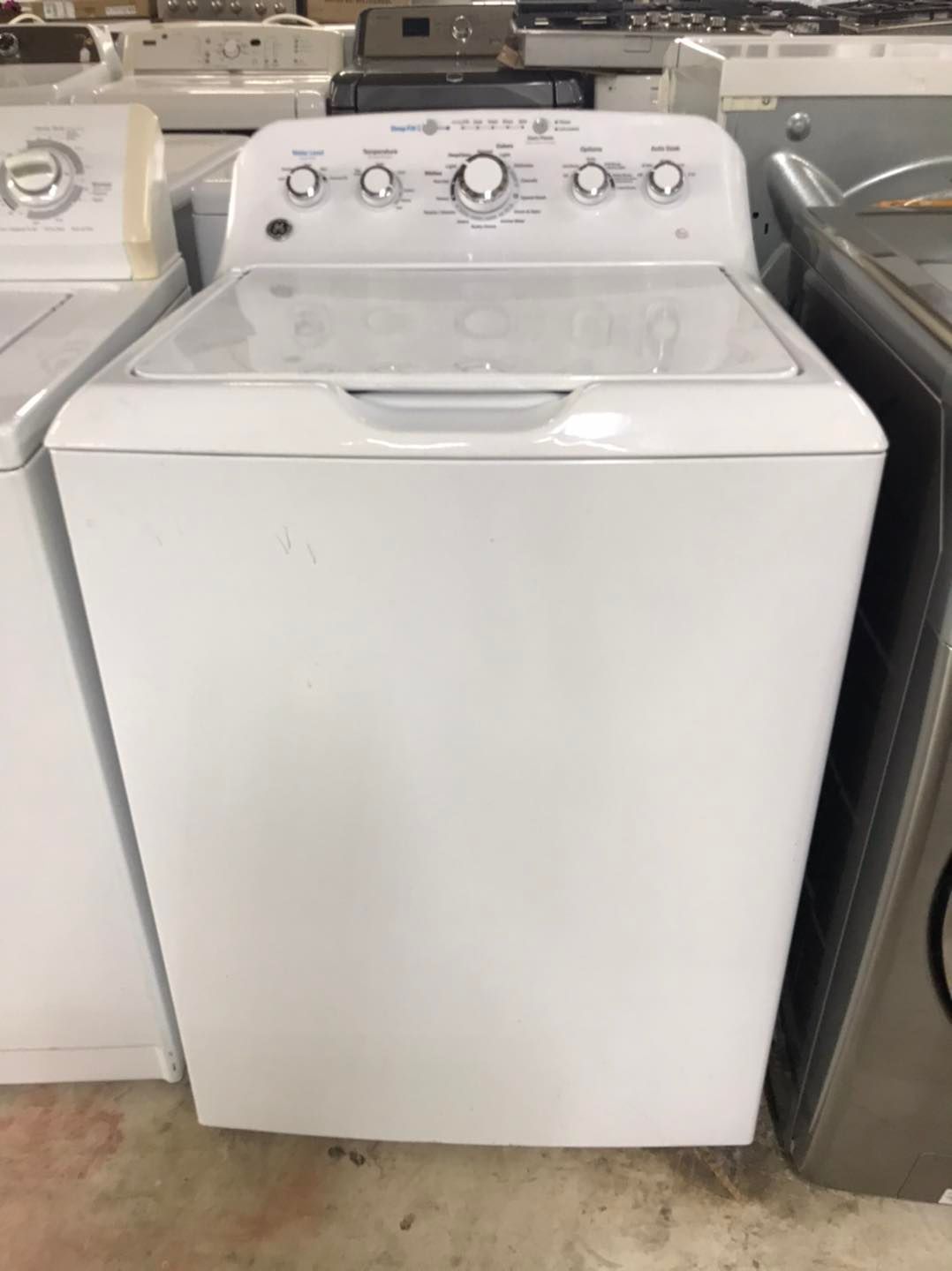 White GE top-load washing machine with knobs on control panel, inside a room.