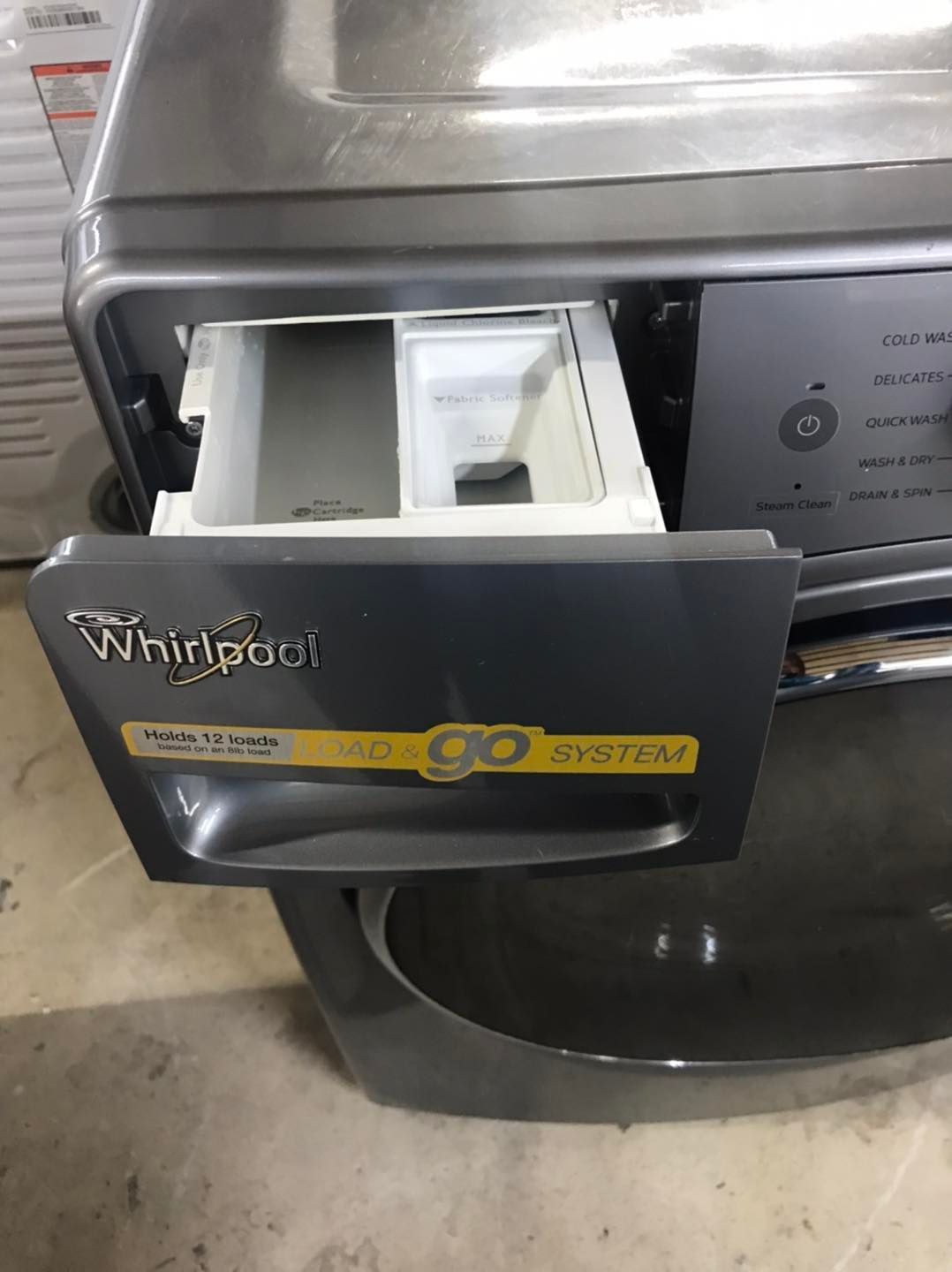 Close-up of a Whirlpool washing machine detergent dispenser drawer, pulled open. Gray machine with white and yellow accents.