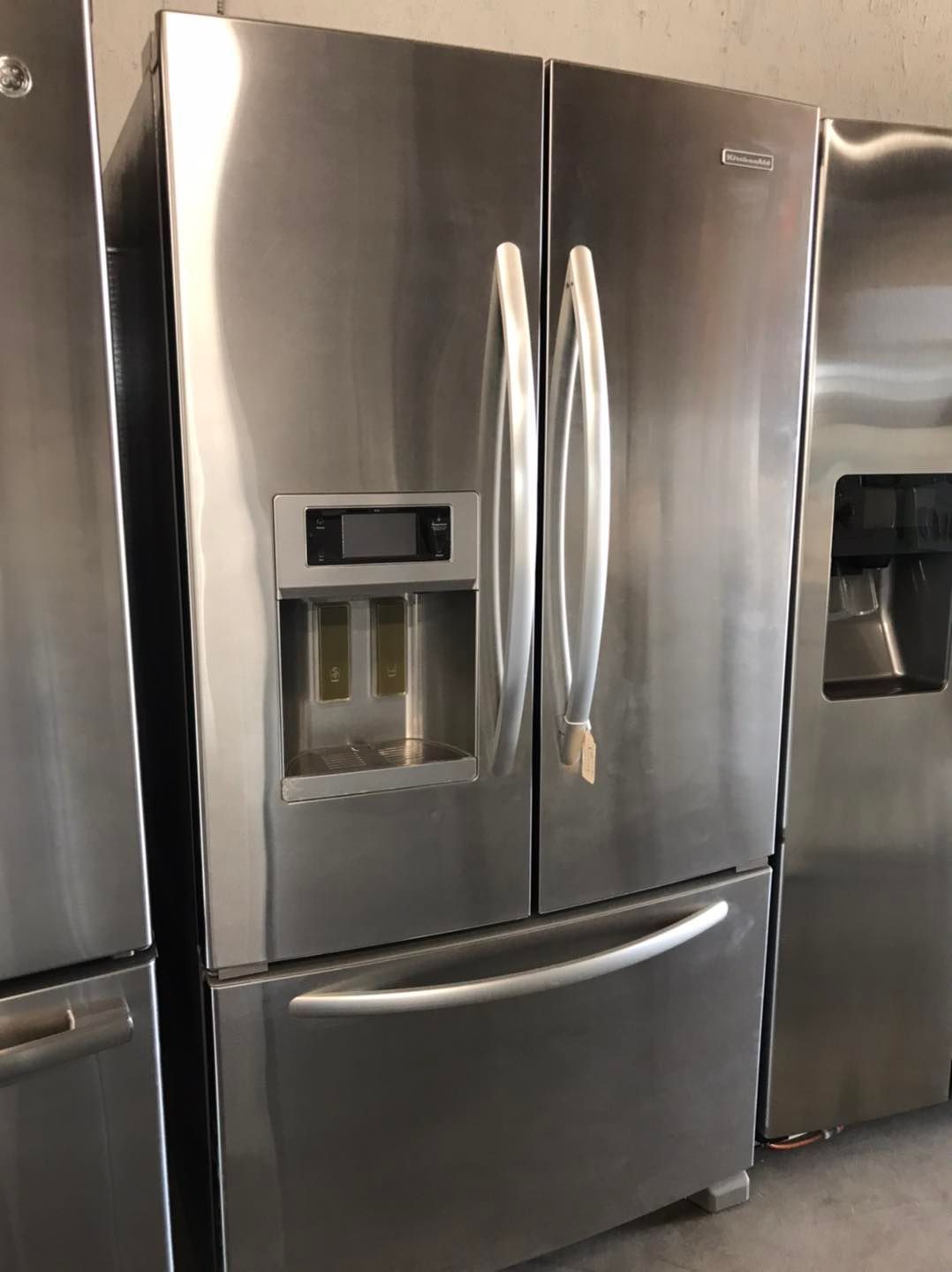 Stainless steel French-door refrigerator with water dispenser and bottom freezer, in a showroom.