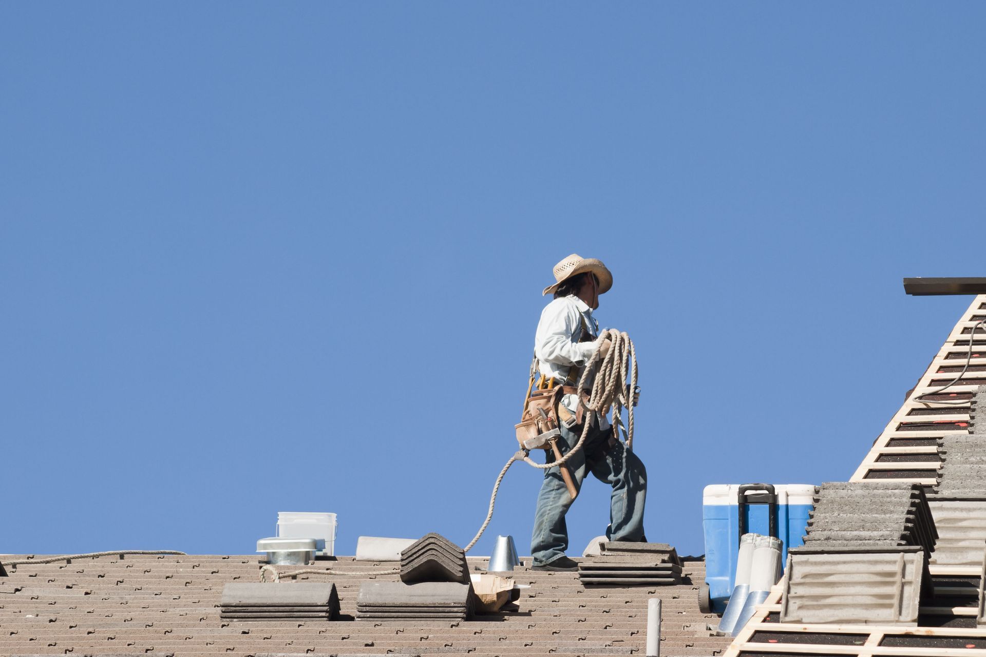 Roofer wearing a hat on a rooftop, holding a rope with a clear blue sky background.