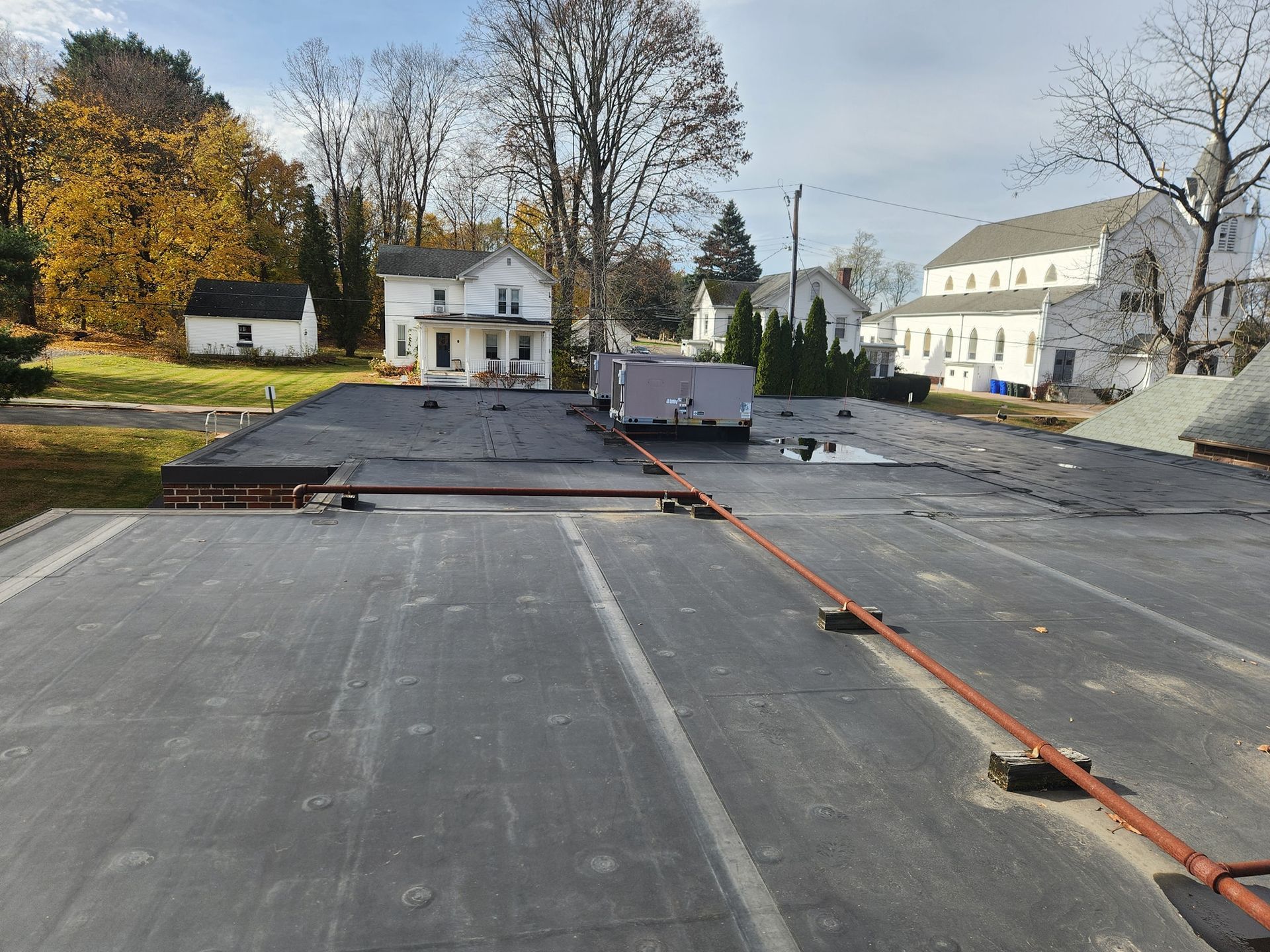 Flat, black roof with red pipes. Buildings and trees in background under a blue sky.