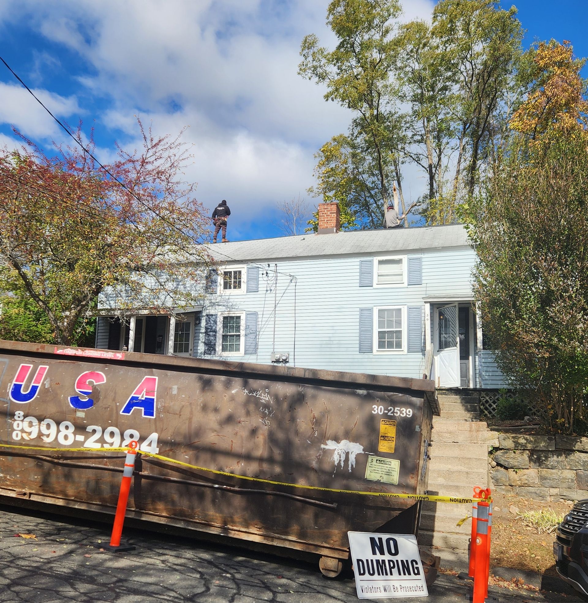 House with blue siding, dumpster in front, person on roof.
